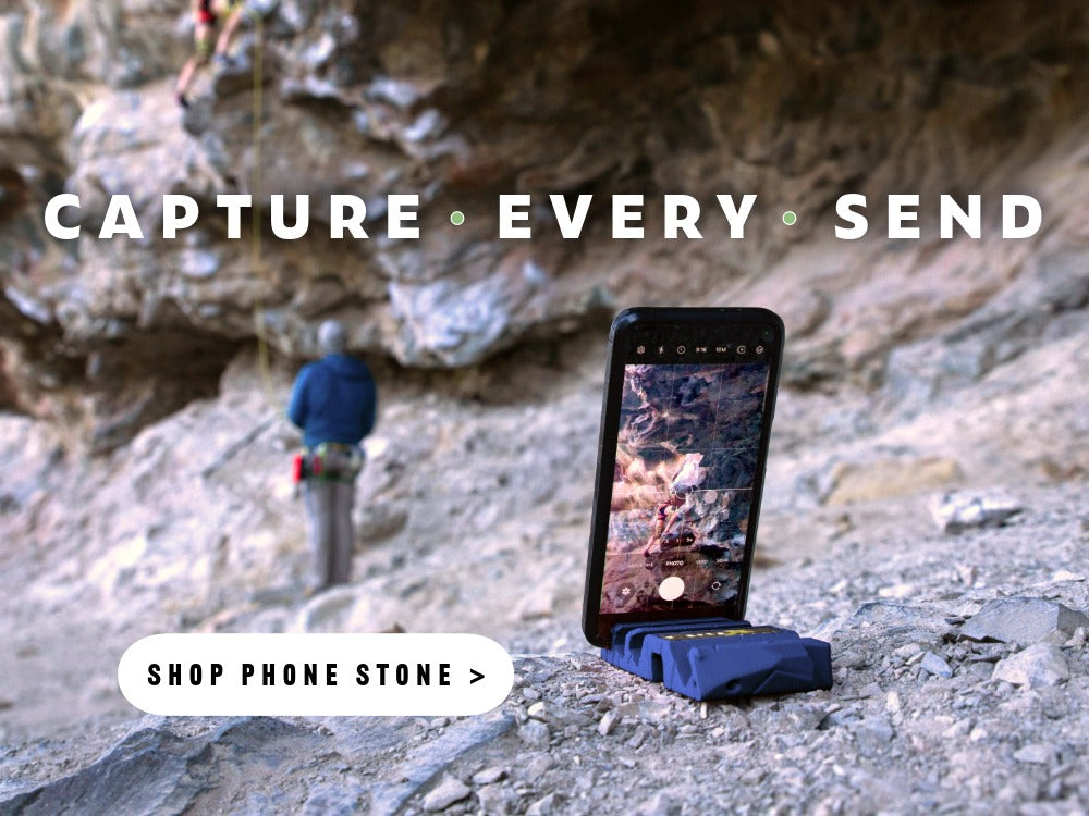 A rock climber climbs outdoors while a belayer stands below. The climbing progress is captured on a mobile phone being held in place by a Crux Gear Phone Stone phone holder.